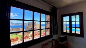 A room with large windows offering a view of a coastal landscape, a bookshelf, and a television on a stand. - Punta Est - PERLA Two-Bedroom Apartment with Terrace and Ocean View (Capo Coda Cavallo)