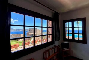 Ein Zimmer mit großen Fenstern und Blick auf eine Küstenlandschaft, einem Bücherregal und einem Fernseher auf einem Schrank.