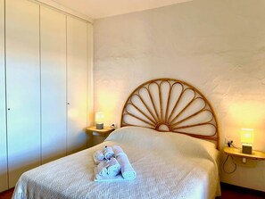 A hotel room with a bed, a wardrobe, two bedside tables with lamps, and a wall-mounted headboard. - Punta Est - PERLA Two-Bedroom Apartment with Terrace and Ocean View (Capo Coda Cavallo)
