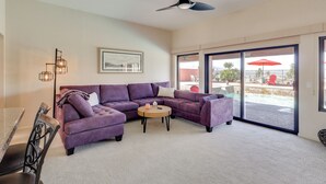 Una sala de estar con un sofá seccional morado, una mesa de centro, un ventilador de techo y un amplio ventanal corredizo que da a un área exterior.