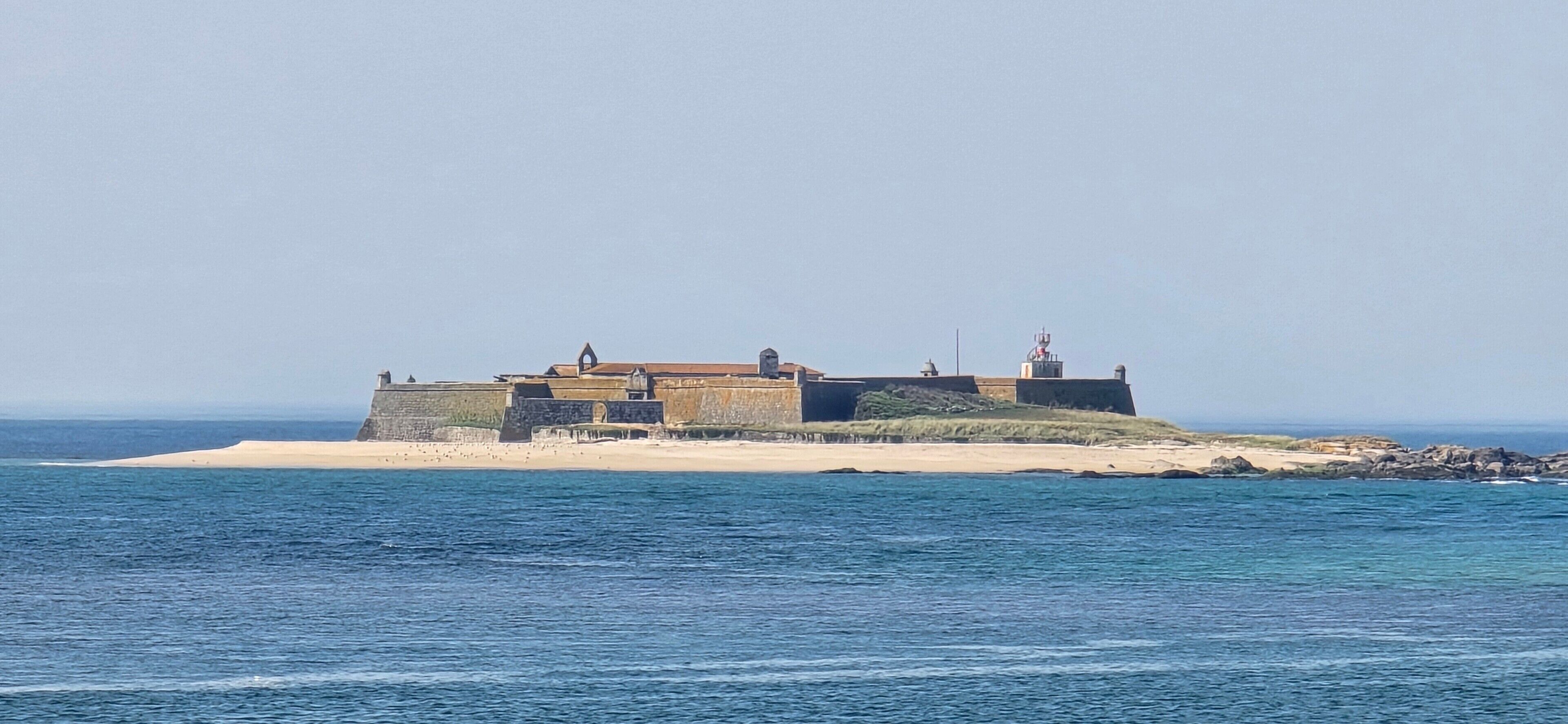 A coastal fortification with a lighthouse, situated on a sandy beach with the sea in the foreground.