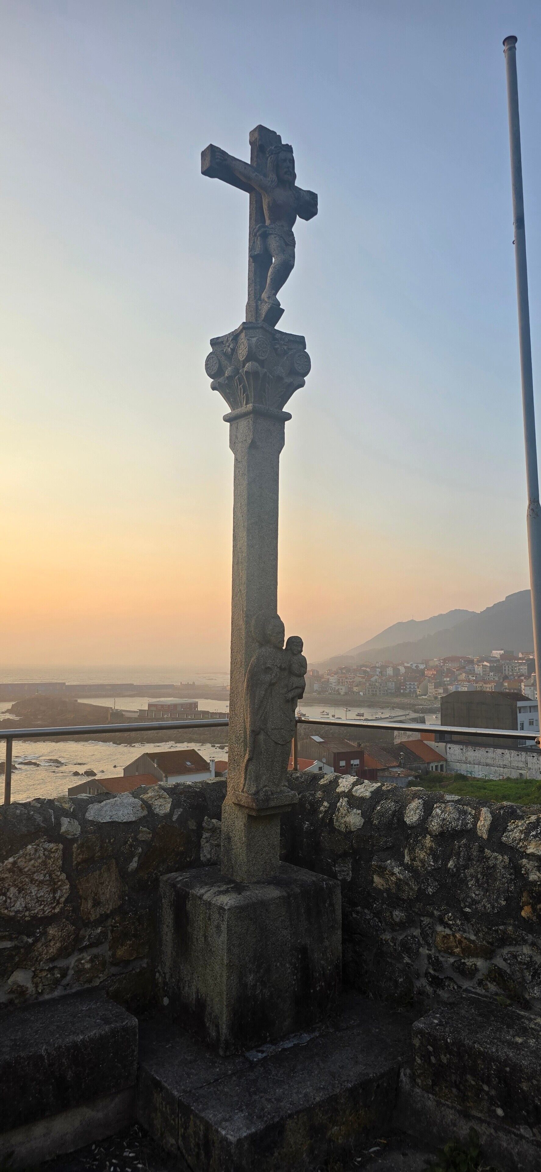 A stone cross with a figure, set against a sunset backdrop with a coastal cityscape.