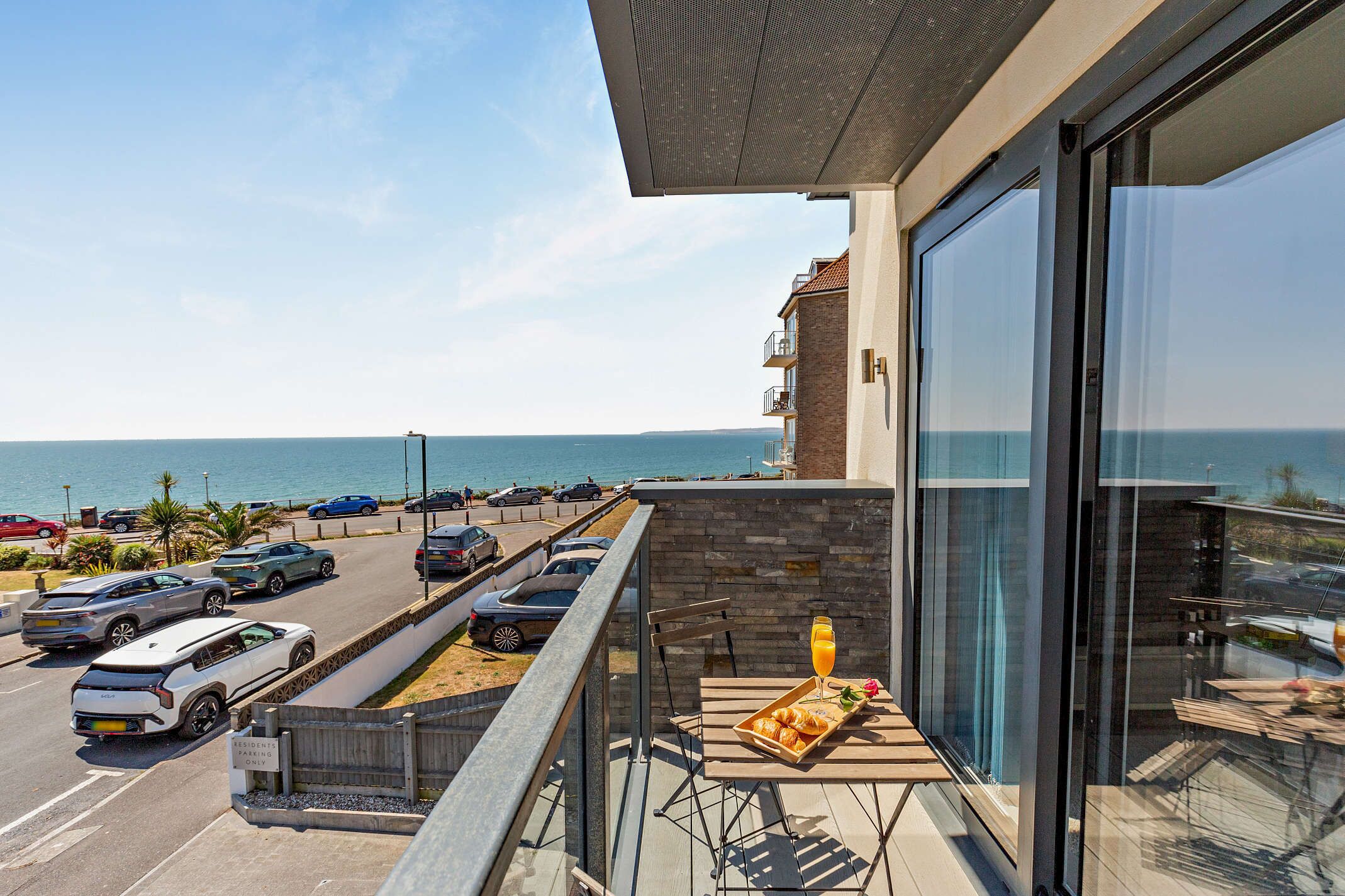 Ein Balkon mit Frühstückstisch, Blick auf das Meer und ein Parkplatz mit Autos.