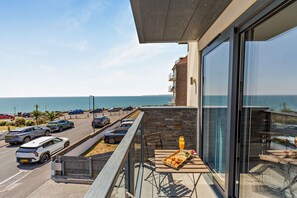 Un balcón con mesa lista para el desayuno, vista al mar y un estacionamiento con coches.