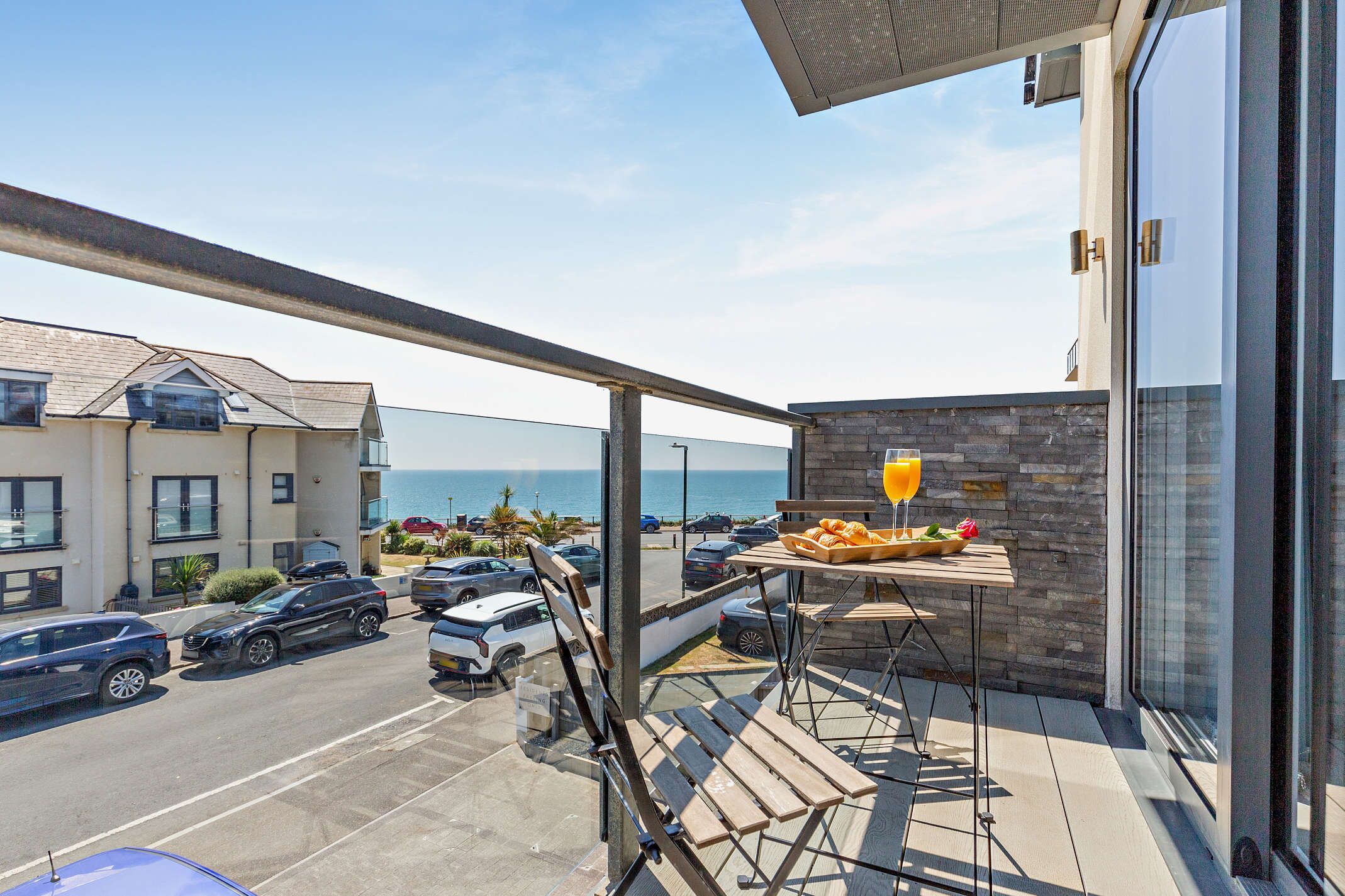 Un balcon avec une table préparée pour le petit-déjeuner, d’où l’on peut admirer un parking et la mer.
