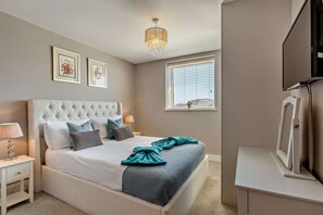 Ein Schlafzimmer mit einer gesteppten Kopfteil, einem Bett mit weißer und gemusterter Bettwäsche, einer Nachttischlampe, einem Fenster mit Jalousien und einem Fernseher auf einem Schrank.