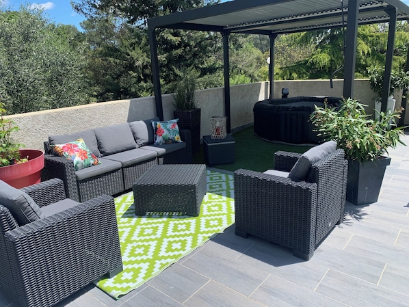 A covered outdoor patio with wicker furniture, a green patterned rug, and potted plants.