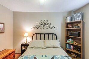 A bedroom with a bed, bedside table, lamp, decorative wall piece, and a wooden shelf with various items.