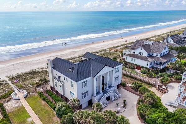 Een groot wit gebouw met een balkon, nabij een strand met een houten looppad.