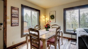 Uma sala de jantar com mesa de madeira, cadeiras e vista para o exterior através de grandes janelas.