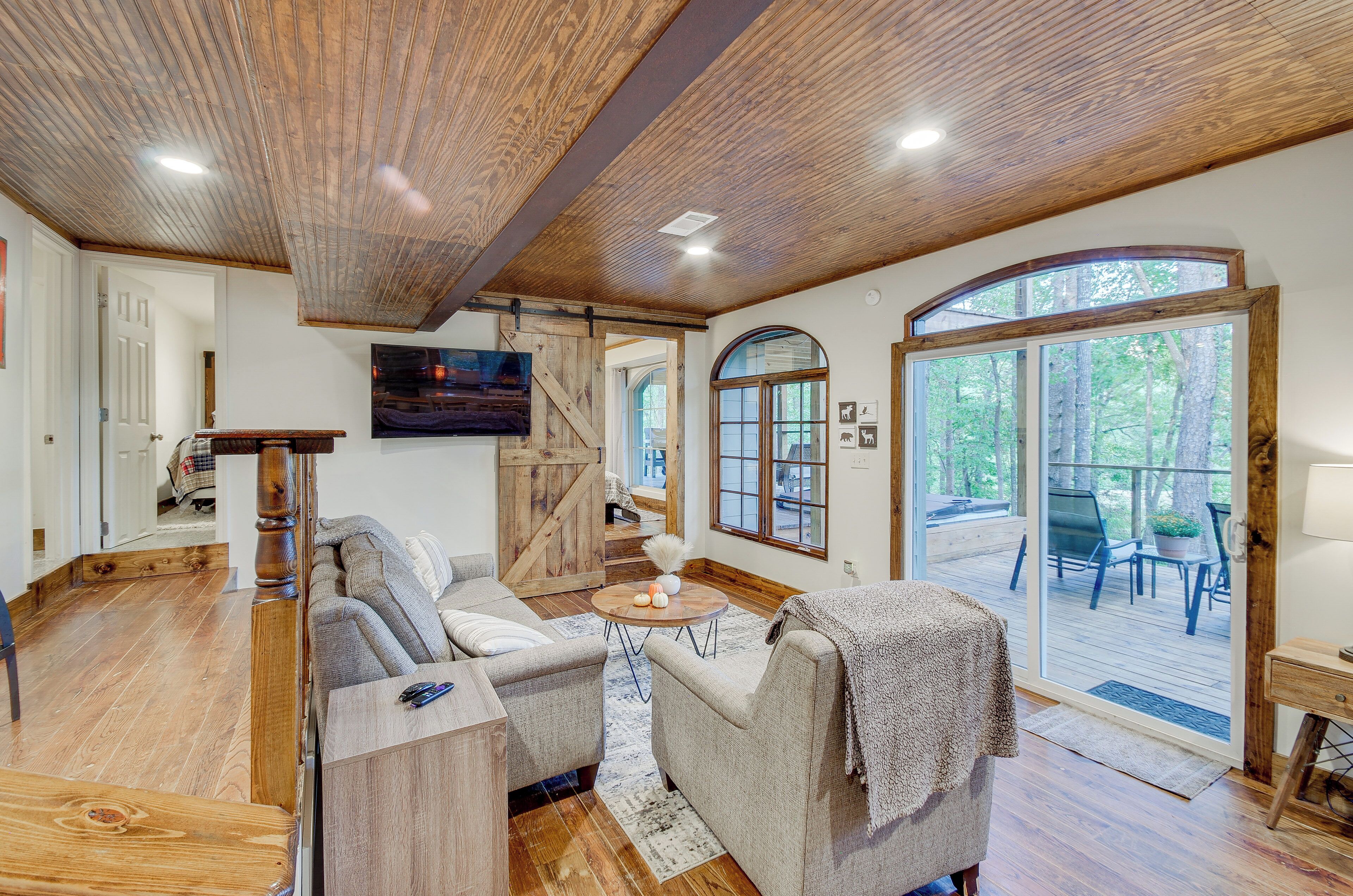 A spacious living room with a wooden ceiling, a large window, and a sliding glass door leading to an outdoor deck.