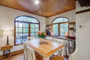 A dining area with a wooden table, white chairs, and a vase of flowers. There are large arched windows with a view of trees.