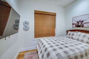Une chambre à coucher avec une couvre-lit à carreaux, une tête de lit en bois, une fenêtre avec des stores et un tableau représentant une vache accroché au mur.