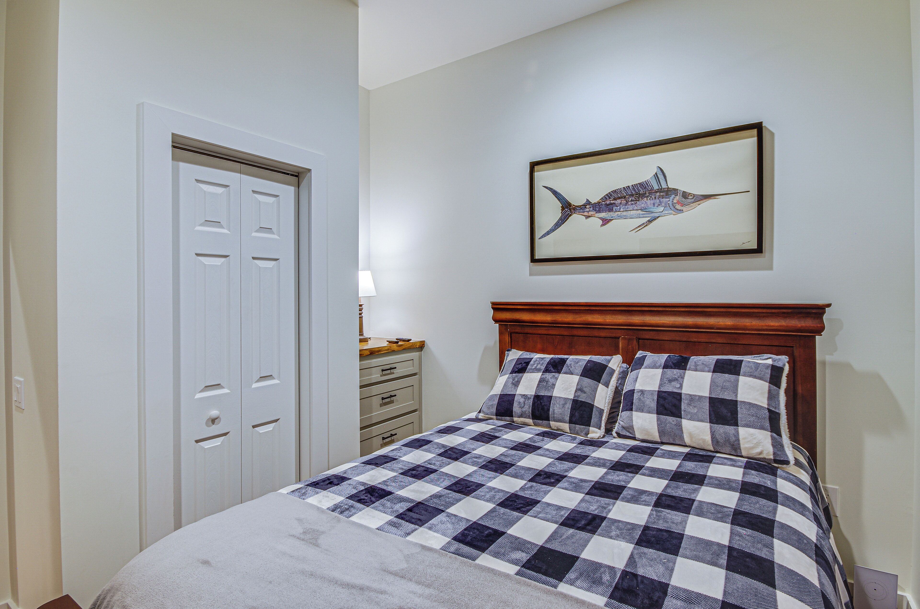 A bedroom with a checkered bedspread, a wooden headboard, a framed fish painting on the wall, and a white door with a mirror.