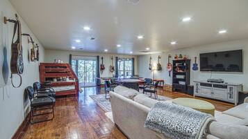 A spacious living room with a wooden floor, a sofa, a dining table with chairs, and a TV mounted on the wall.