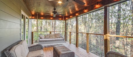 Un espacio de estar al aire libre techado con muebles de mimbre, ventilador de techo y vistas al bosque.