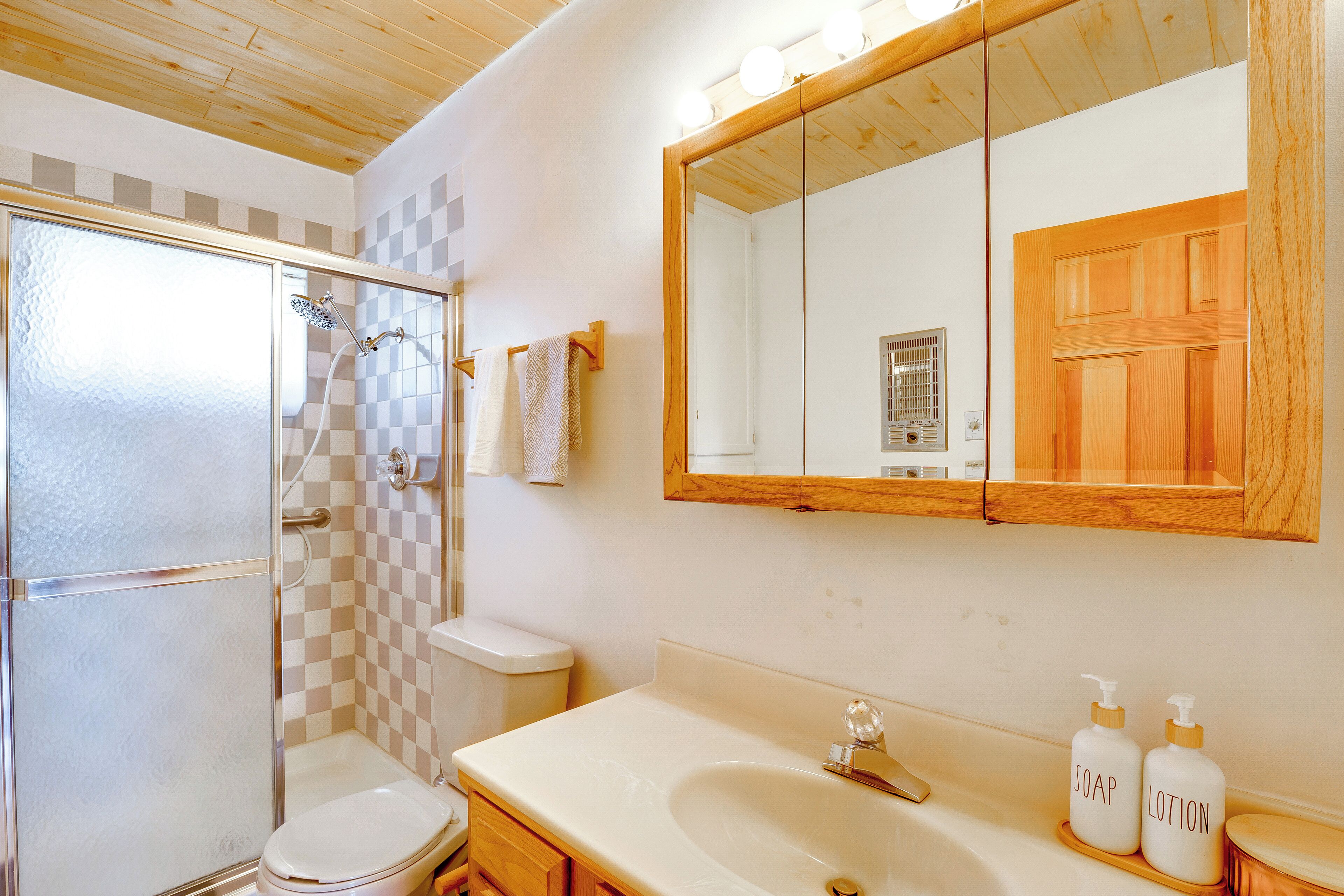 Une salle de bain avec une douche à parois de verre, des toilettes, un lavabo avec des flacons de savon et de lotion, et un miroir avec un cadre en bois.