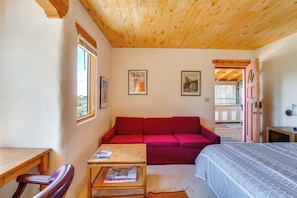 A cozy room with a wooden ceiling, a red sofa, a bed with a patterned bedspread, a small table with a magazine, and a window with curtains. - Des Montes Retreat: Hike, Ski, Swim & Shop! (El Prado)