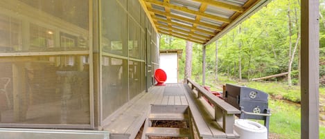 A covered outdoor area with a wooden bench, a grill, and a large window overlooking a forested area.