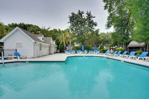 A swimming pool with blue lounge chairs and umbrellas, surrounded by trees and a white building. - Charleston Townhome Near Credit One Stadium (Charleston)