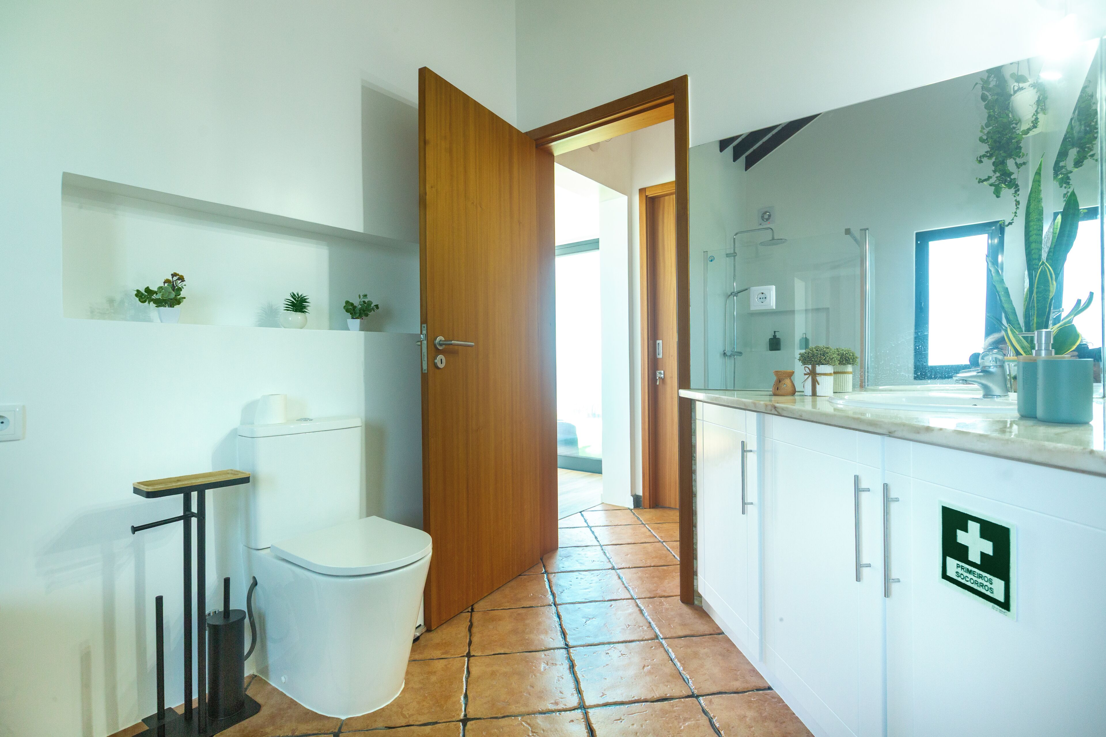 A bathroom with a toilet, a sink, and a shower. There are potted plants on the shelves and a sign indicating first aid.
