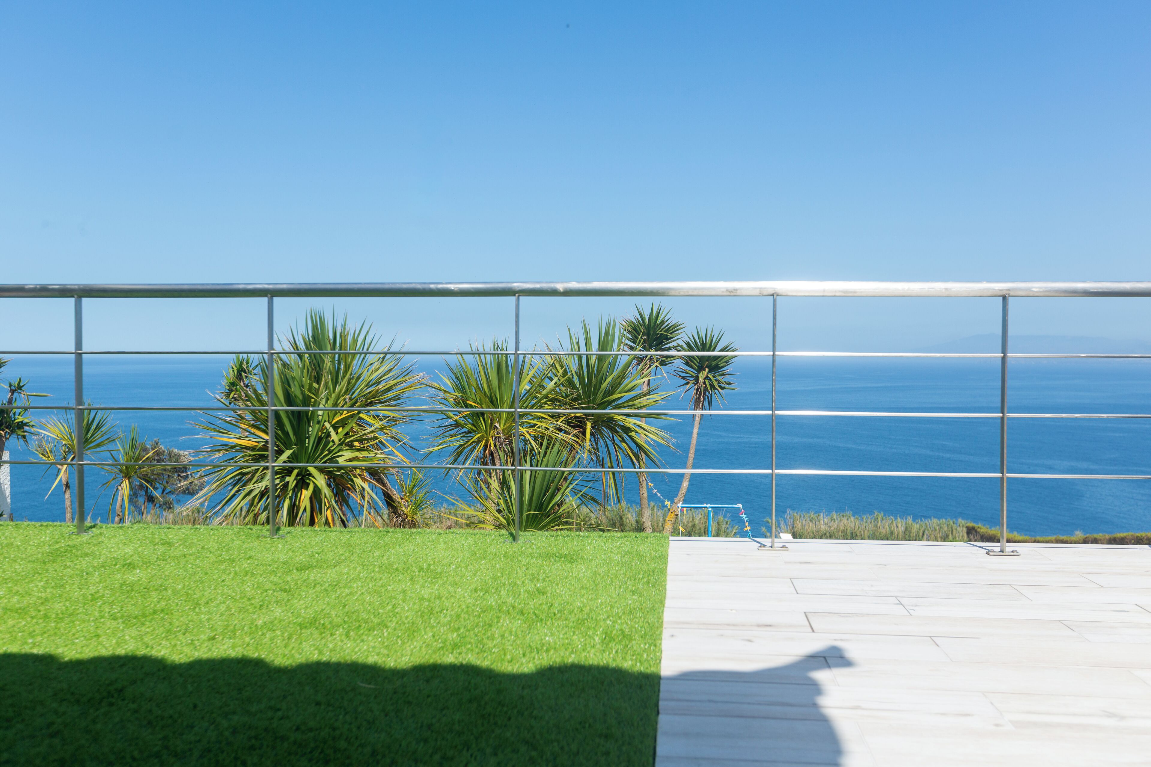 A balcony with a clear view of the ocean, a grassy area, and a metal railing.