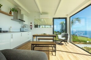 A modern kitchen with white cabinets, a wooden dining table, and a view of the ocean through large windows. - Costa Bela Retreat Azores: The perfect getaway, offering comfort, nature, and serenity. (Açores)