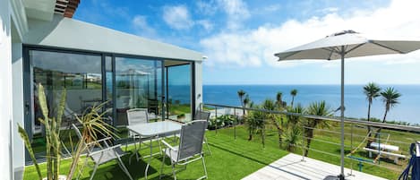 A modern outdoor patio with a glass enclosure, a dining area, and a view of the ocean.