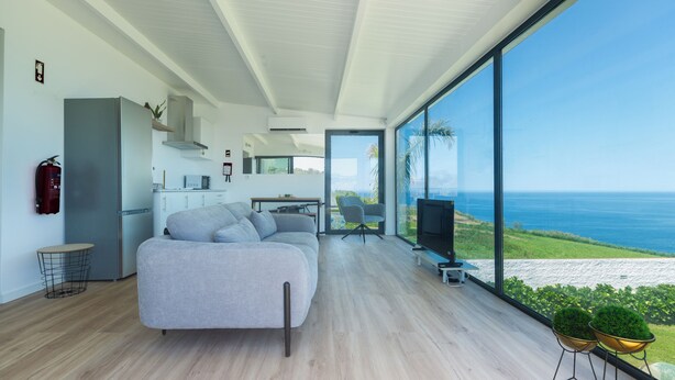 A modern living room with a large grey sofa, a flat-screen TV, and a view of the sea. - Costa Bela Retreat Azores: The perfect getaway, offering comfort, nature, and serenity. (Açores)