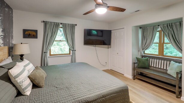 A bedroom with a bed, a bench, a ceiling fan, a TV, and a window with curtains. - Mins from AVL, Woodsy Weaverville Retreat (Weaverville)