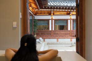 En traditionell japansk inredning med träbjälkar, tatamimattor på golvet och utsikt över en innergård med en stenlykta.