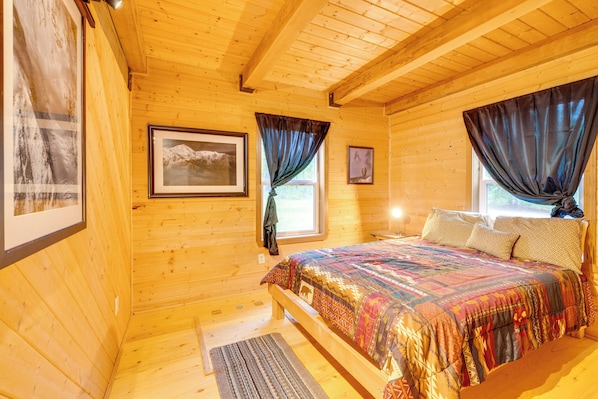 Ett sovrum i en stuga med träpanel, en säng, inramade bilder och ett fönster med gardiner.