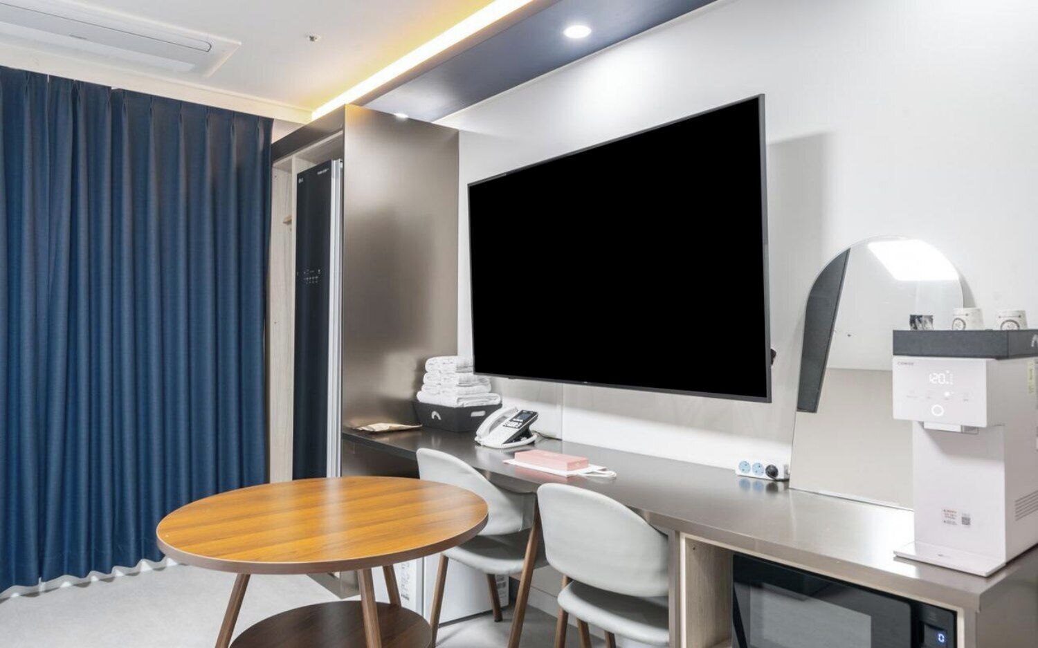 A hotel room with a large flat-screen TV, a desk with a telephone, and a chair.