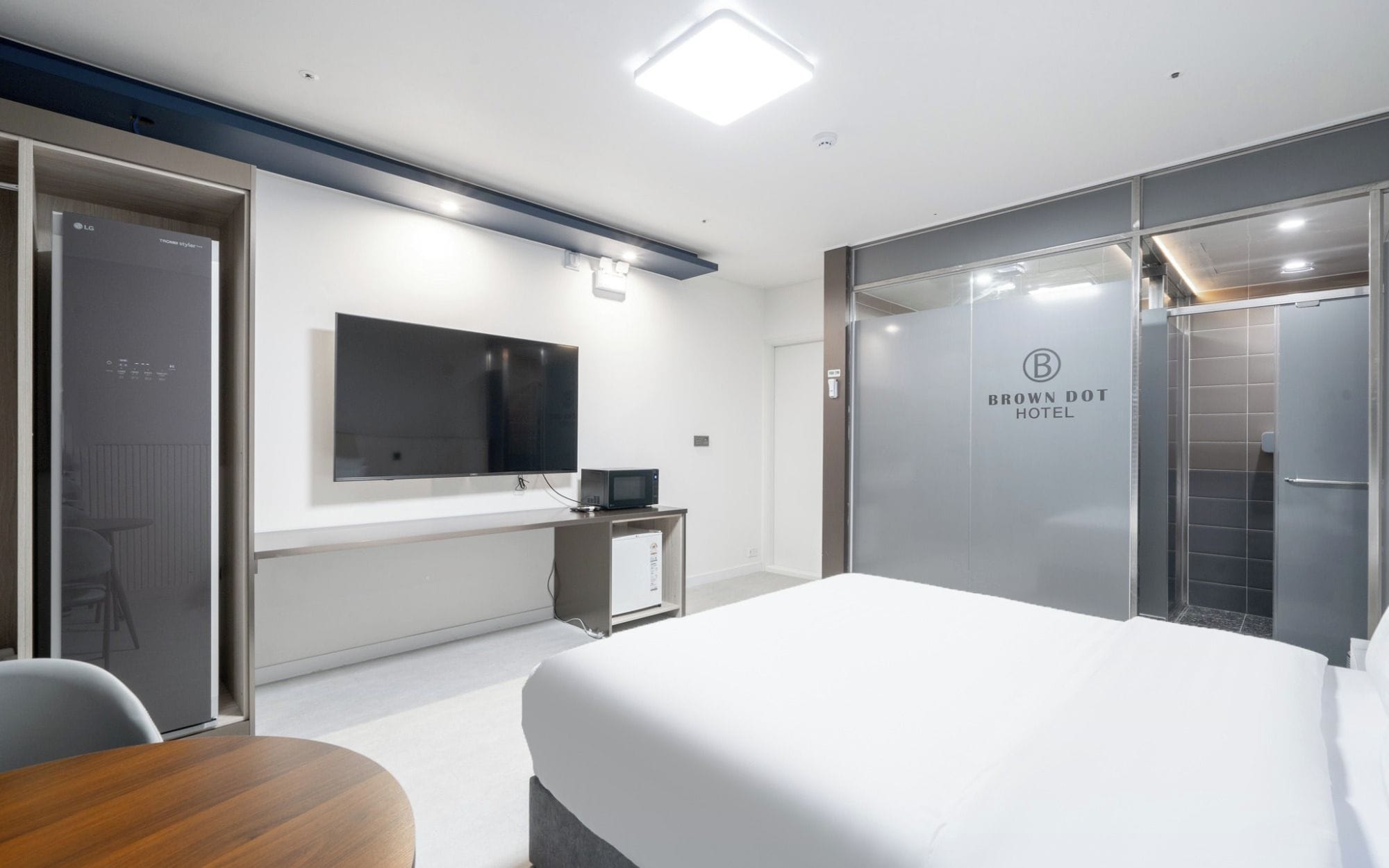 A modern hotel room with a large bed, a flat-screen TV mounted on the wall, a small desk with a computer, and a glass partition with the hotel name.