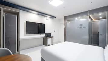 A modern hotel room with a large bed, a flat-screen TV mounted on the wall, a small desk with a computer, and a glass partition with the hotel name.