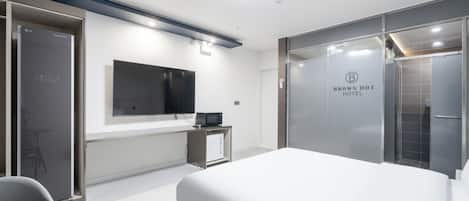 A modern hotel room with a large bed, a flat-screen TV mounted on the wall, a small desk with a computer, and a glass partition with the hotel name.