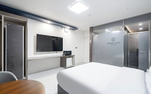 Habitación de hotel moderna con una cama grande, un televisor de pantalla plana montado en la pared, un escritorio pequeño con una computadora y un panel de vidrio con el nombre del hotel.