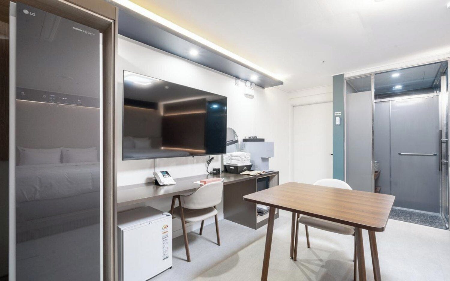 A hotel room with a bed, a desk, a chair, a flat-screen TV, a mini-fridge, and a shower area.