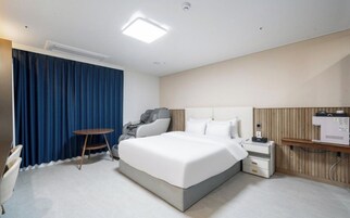 A hotel room with a bed, a massage chair, a small table, a nightstand, and a wall-mounted heater.