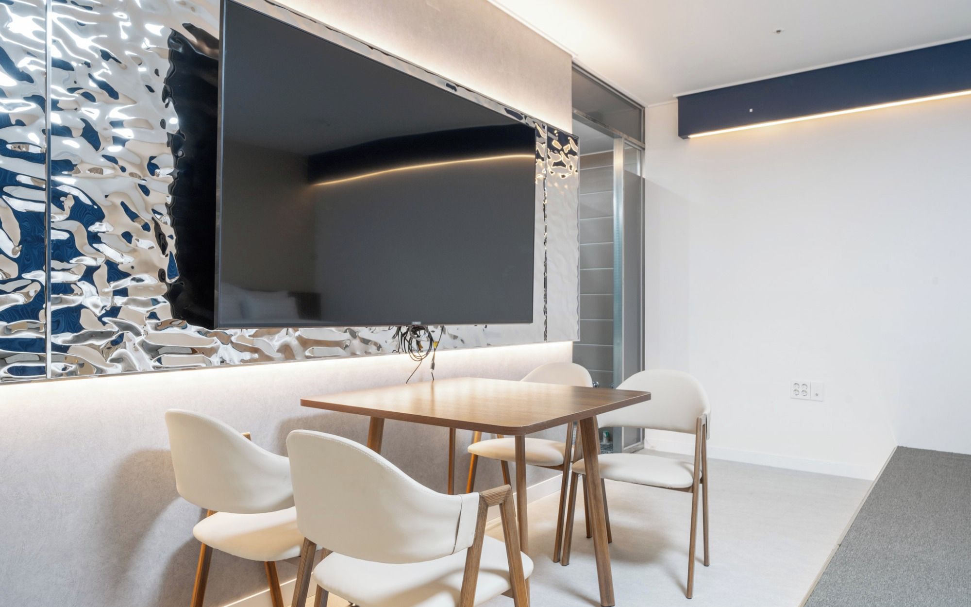 A modern meeting room with a large flat-screen TV, white chairs, and a wooden table.
