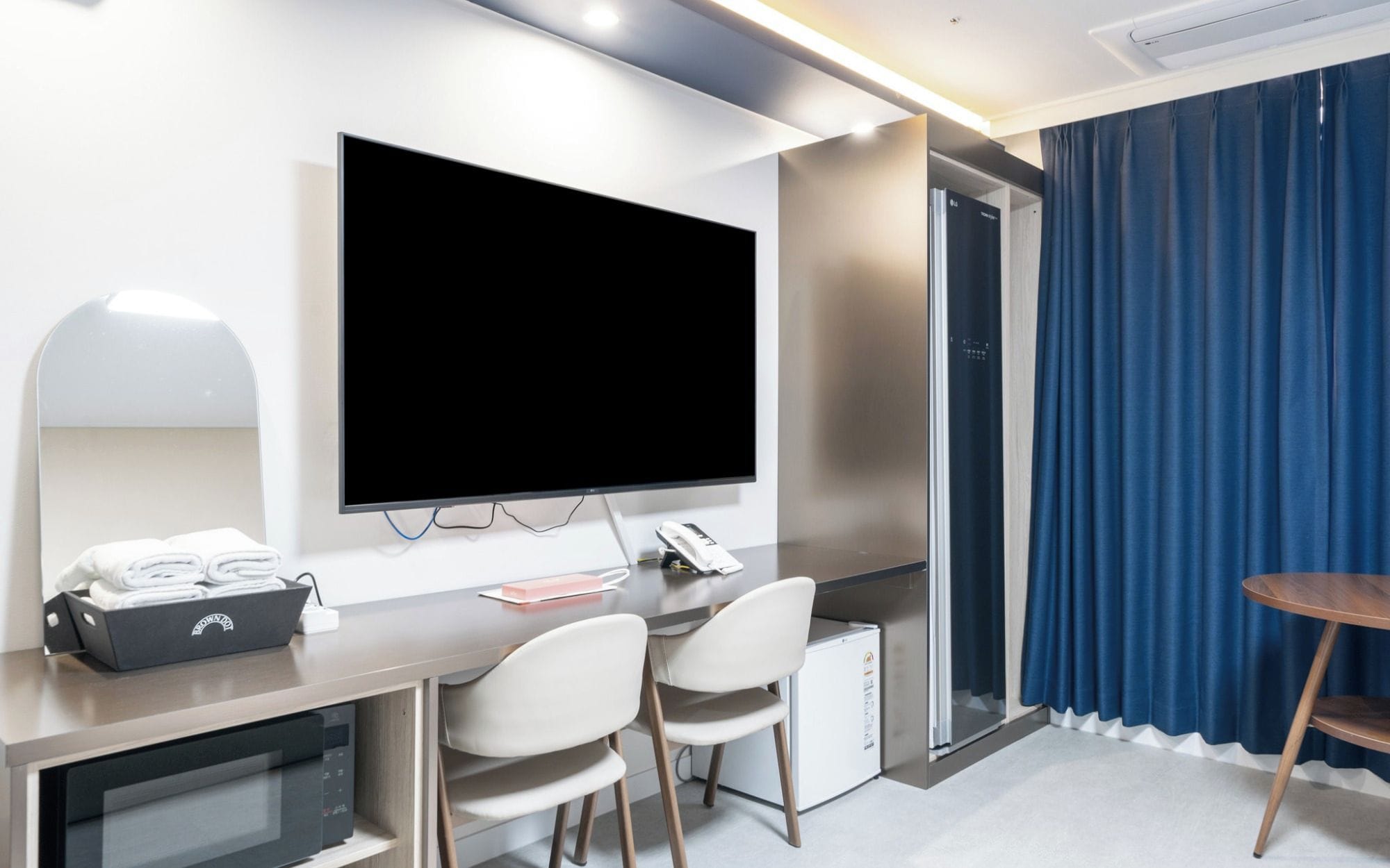 Quarto de hotel com televisão de tela plana na parede, uma escrivaninha com telefone, um pequeno frigorífico e duas cadeiras.