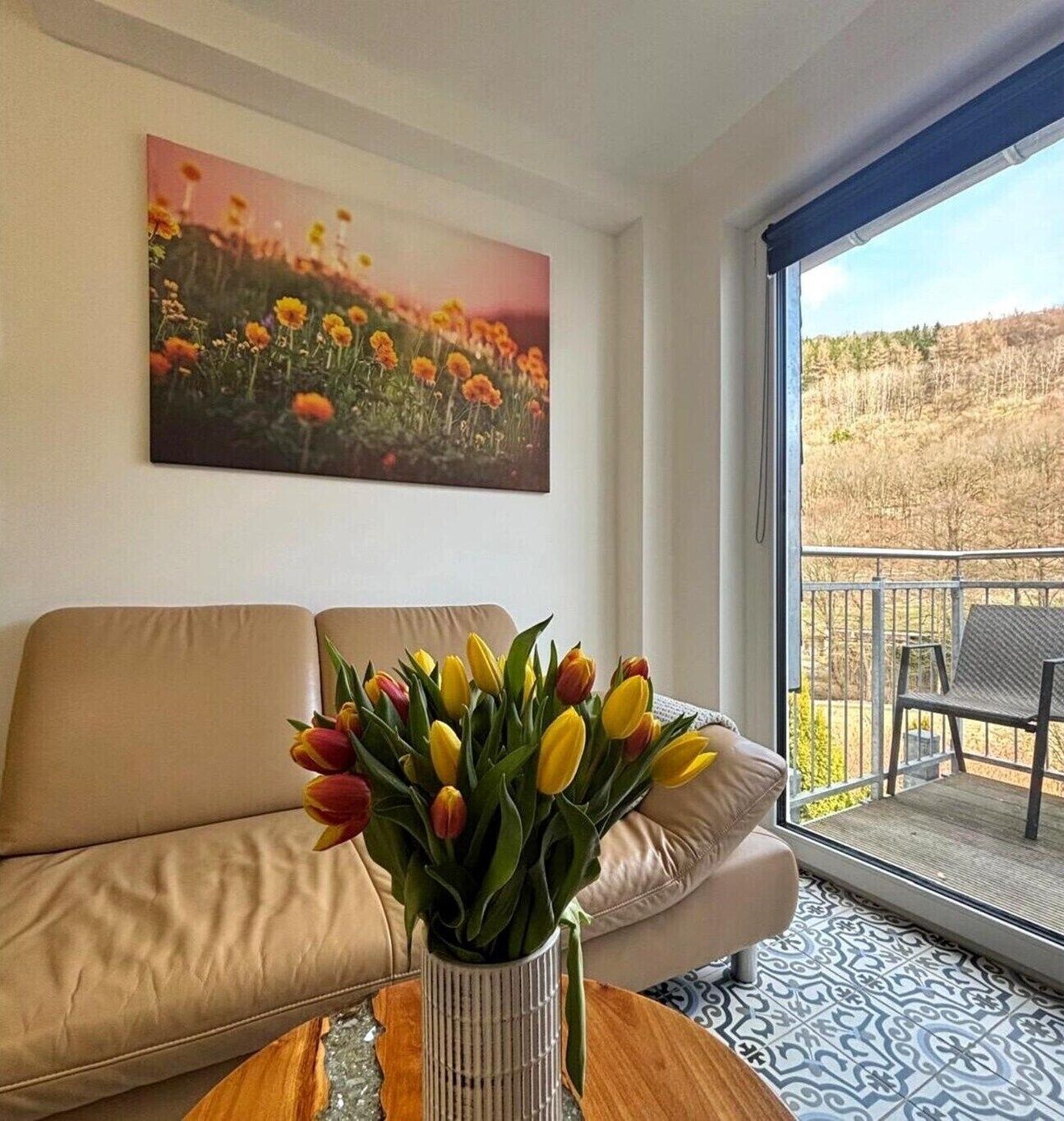 Een woonkamer met een bank, een bijzettafeltje met een vaas tulpen, een schilderij aan de muur en uitzicht door een glazen deur.