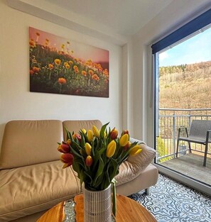 A living room with a sofa, a coffee table with a vase of tulips, a painting on the wall, and a view through a glass door. - Mehrere Ferienwohnungen in Willingen (Willingen)