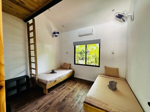Ein kleiner, kompakter Raum mit zwei Einzelbetten, einer Holzleiter, einem Fenster mit Blick auf Bäume und einem Wandventilator.