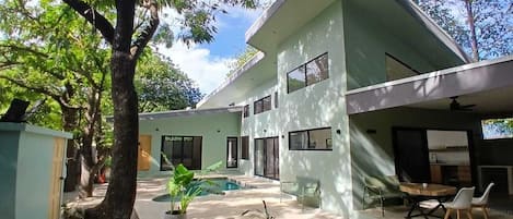 A modern two-story house with a swimming pool, outdoor seating, and a large tree.