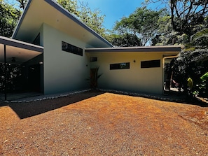 Una casa moderna con un patio cubierto, camino de entrada de grava y un pequeño jardín con una palmera.