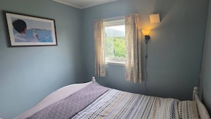 A bedroom with a bed, a window with curtains, a wall-mounted lamp, and a framed picture.
