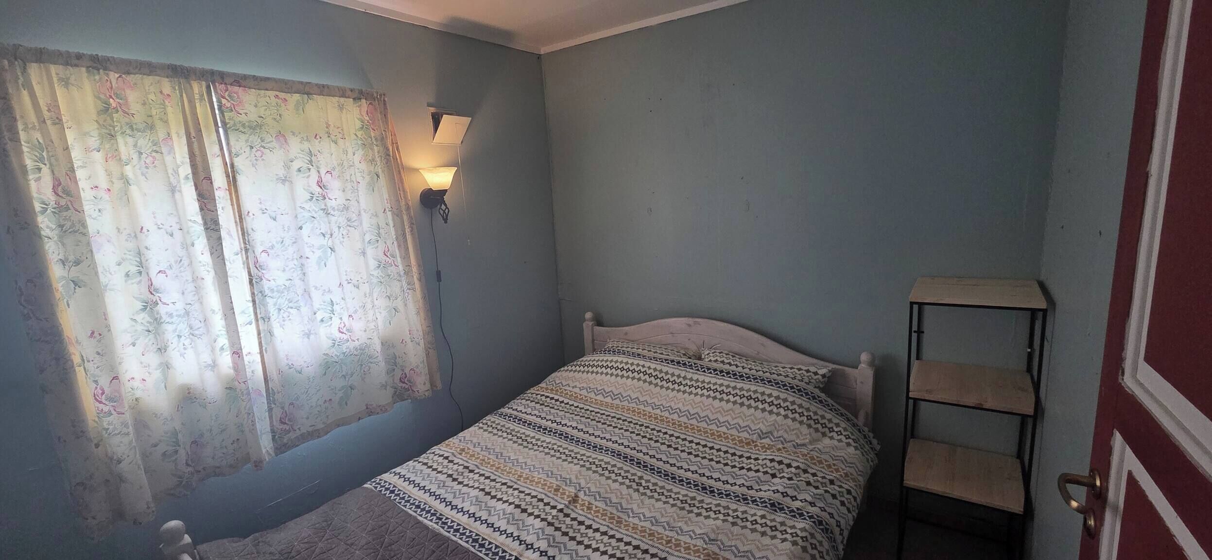 A bedroom with a single bed, a nightstand, a lamp, and a window with floral curtains.