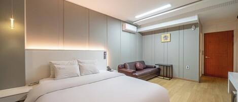 A hotel room with a bed, a sofa, a bedside table, an air conditioning unit, and a door.