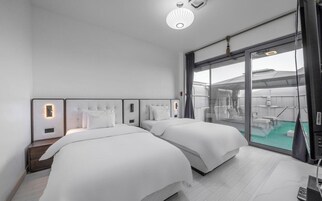 A modern hotel room with two beds, a large window with a view of the outside, and a ceiling light fixture.